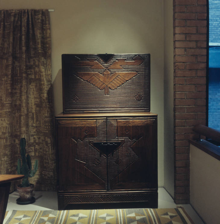 front view of wooden carved desk unit. Top is a drop leaf desk and on the door is carved a symmetrical bird symbol in bold angular style. Bottom portion of desk has two doors - each door has a kneeling figure carved on it. They are down in an angular style and face each other, a man on the left and a woman on the right.