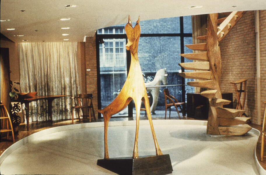 Wood sculpture of a deer stands in the foreground of a gallery space. A Wooden spiral staircase stands to the right, in front of a brick wall, and the back wall of the room is all glass. Through the window is another wood animal sculpture, this one a horse. Inside to the left is a captains chair and a table with a carved bowl on top.