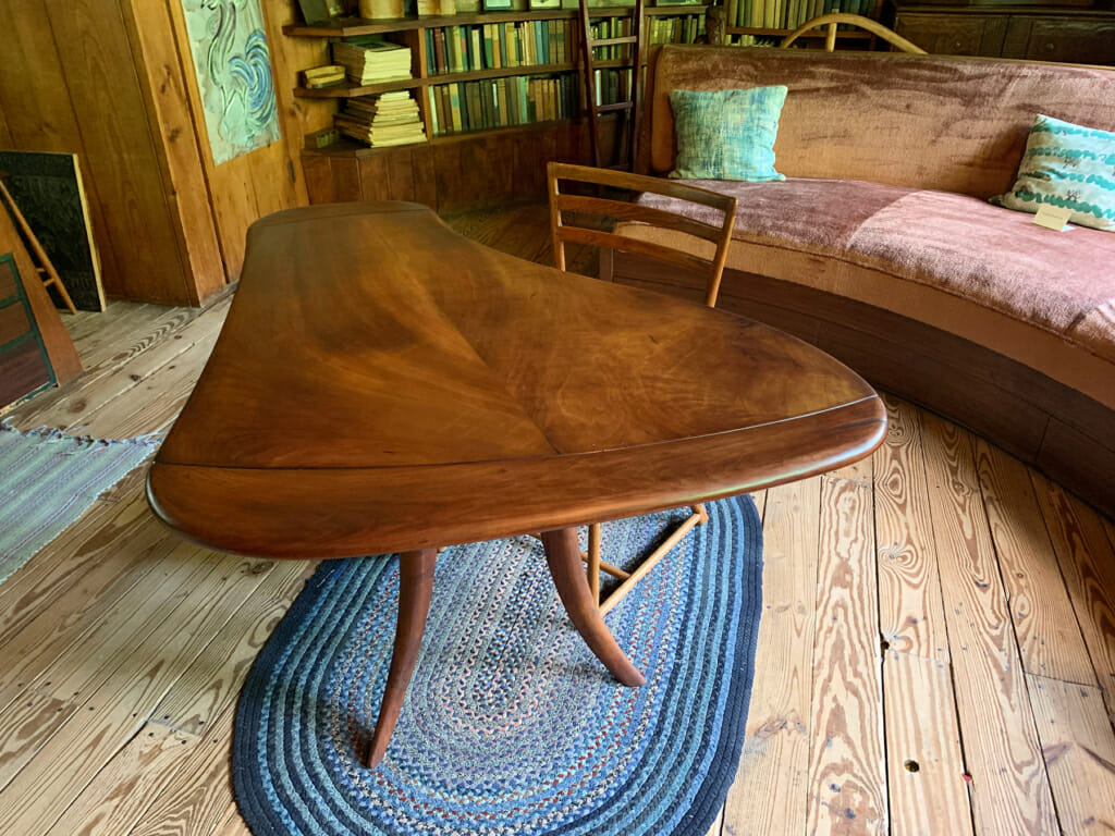 view of Esherick's bedroom showing wooden table with freeform curving top, similar to a grand piano shape. below the table is a small blue braided rug. the is alsoa chair behind the table, a curving sofa and bookshelves at the far end of the room.