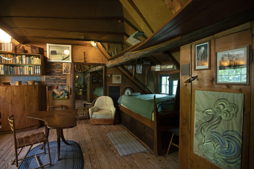 a view of Esherick's bedroom shows bad to the right with light from the window above it. Pictures and drawings hang on the wall and a large mirror hangs on the far side of the room. A leather chair sits near the foot of the bed and a three-legged table with a grand piano-ish shaped top and a chair sit in the center of the room on a blue oval braided rug. Book shelves can be seen above the bed and above a dresser on the far side of the room.