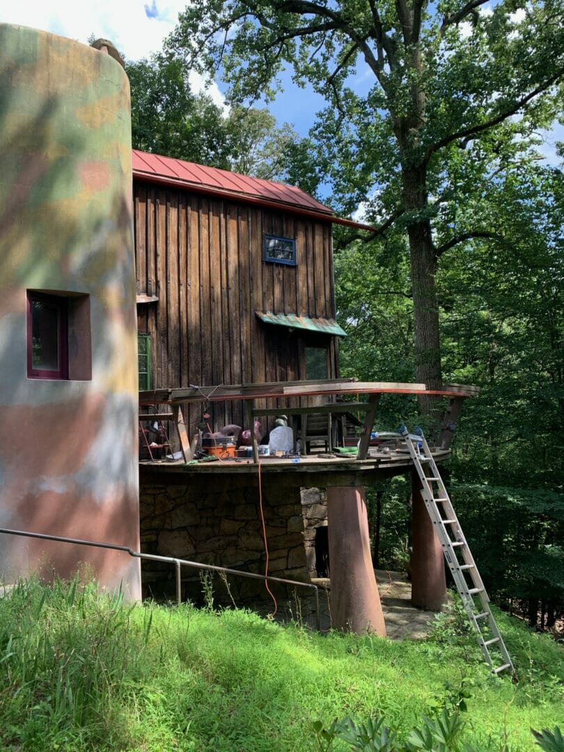 Image shows curving free-form deck on the Studio with grassy area in foreground and a colorful curving stucco wall to the left. A ladder leans up to the deck and tools are strewn about the deck.