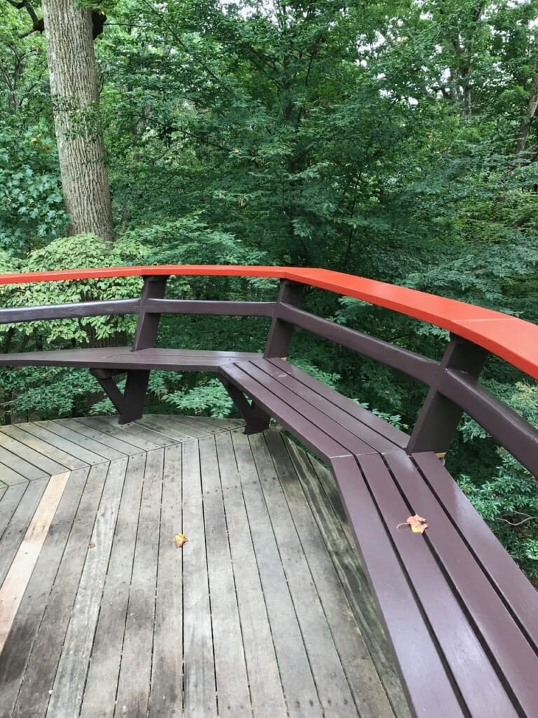 view of deck shows curving red railing and bench with woods in the background