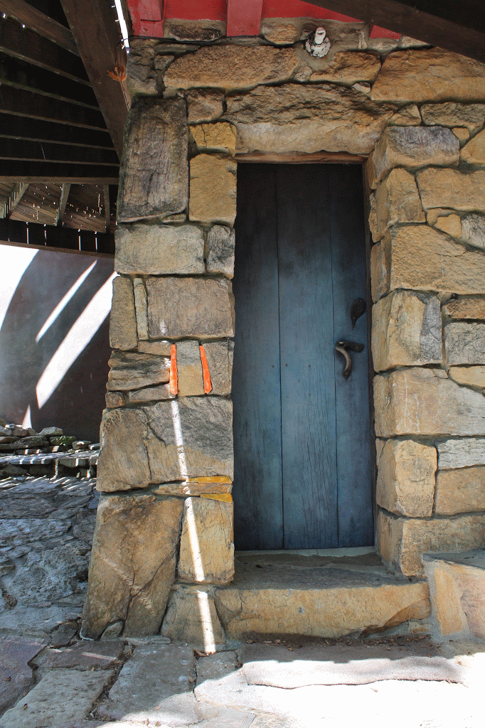 A blue door set in a stone wall is under a deck. Orange and yellow ceramic pieces fir the the cracks in the stone wall. A streak of light walls across the wall
