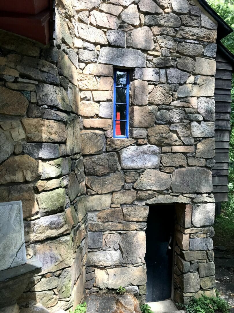 A stone wall with dappled light has a door in the lower right and a tall narrow, blue window in the center. In the window we can see a bright red abstract sculpture.