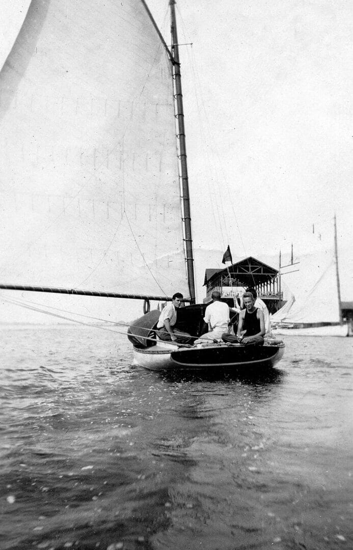 black and white photo of a young Wharton sitting on the back of a sailboat, and facing the viewer. His brothers are also on the boat, which is moving away from us.