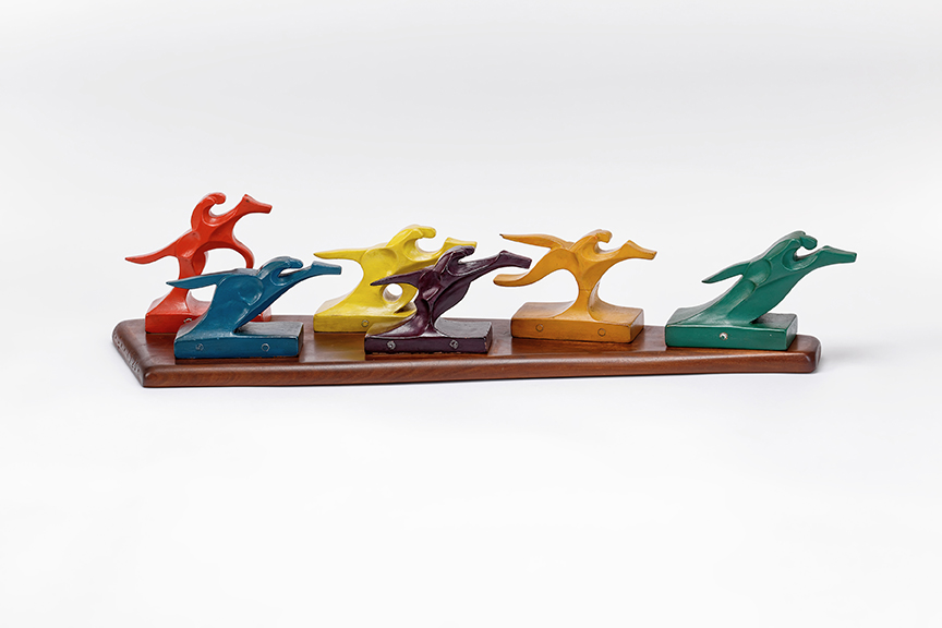 six wooden carved jockeys on horseback mounted to a wood board. The jockeys are painted a variety of different colors.
